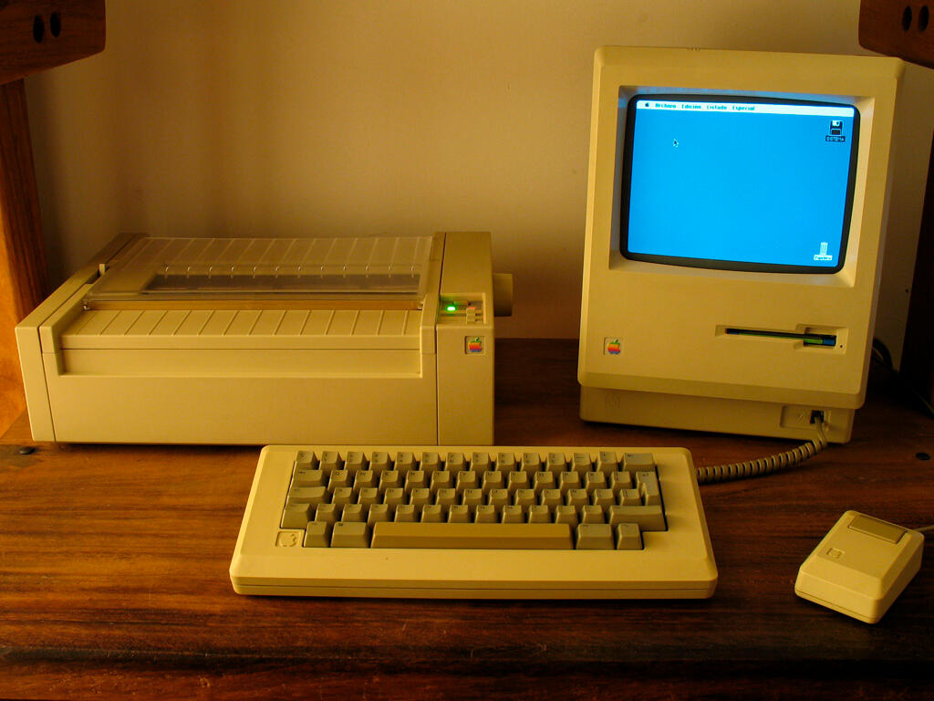Ordenador Apple Macintosh 512K de 1986 en un escritorio de madera, junto a teclado, ratón y una impresora de matriz de puntos.