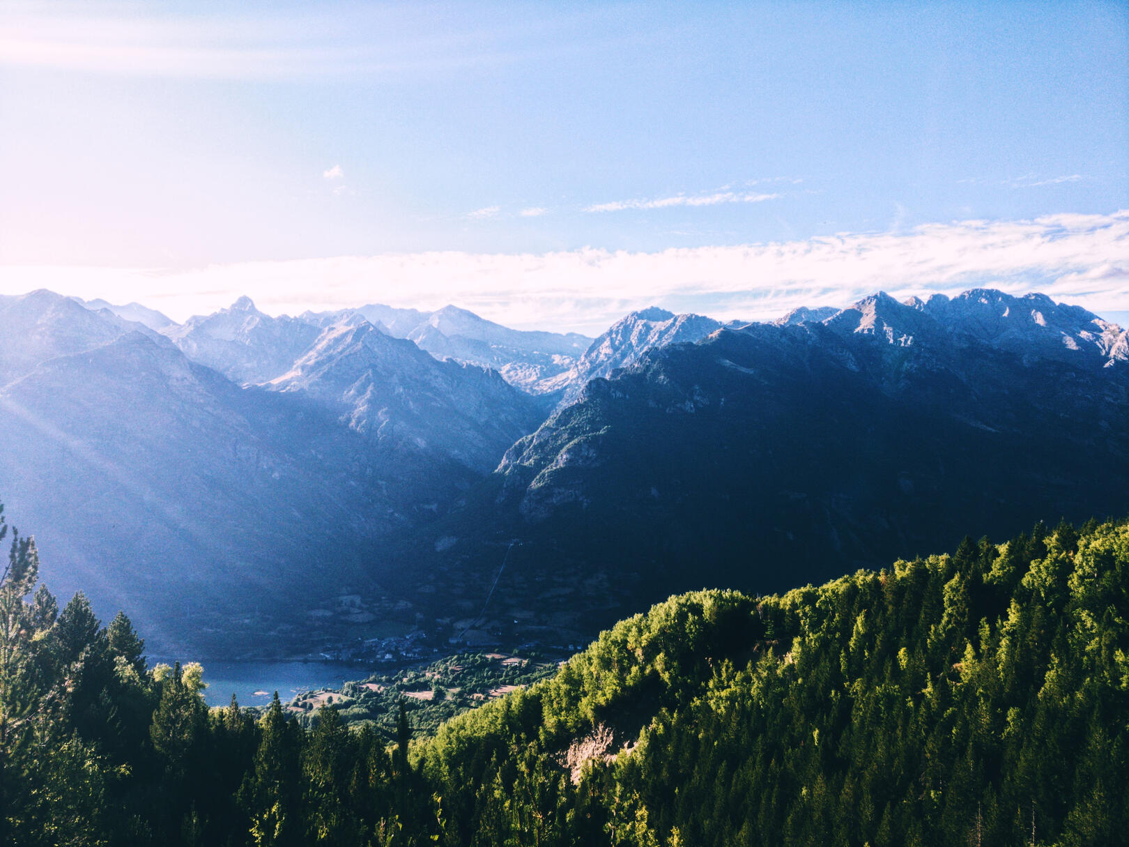 Vista panorámica del valle de Benasque al atardecer, con montañas del Pirineo aragonés al fondo, bosques verdes en primer plano y nubes reposando sobre las cumbres.